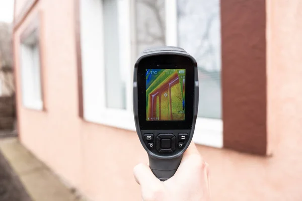 Een mannelijke hand houdt een warmtebeeldcamera bij het raam van een huis. Zoek naar warmteverlies in particuliere huizen — Stockfoto