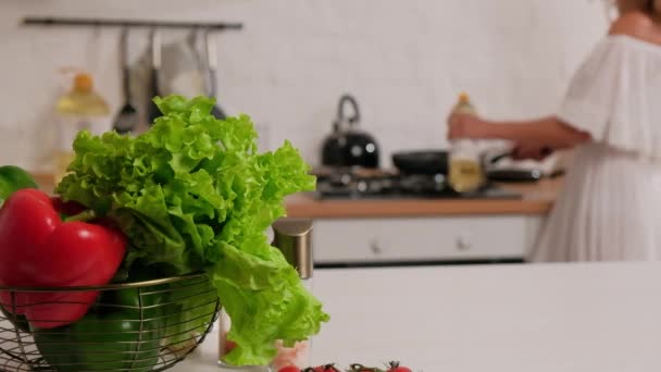 Een vrouw kookt en giet olie in een koekenpan op de achtergrond. Voorgrond verse groenten en tomaten. — Stockvideo
