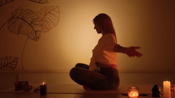 Blond meisje in de lotuspositie in de schemering tijdens het branden van kaarsen op het tapijt. Yoga praktijk en totale ontspanning. — Stockvideo