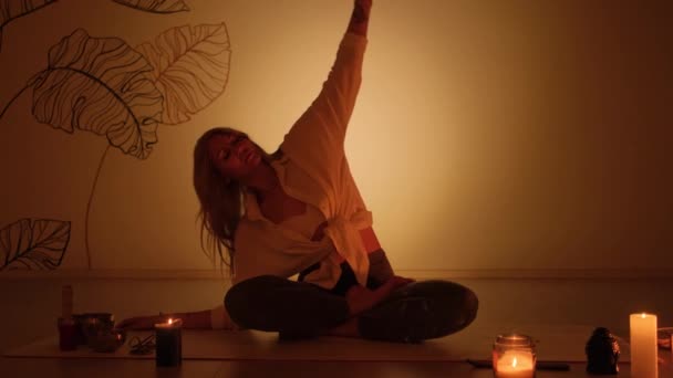 Blondynka w pozycji lotosu o zmierzchu podczas palenia świec na dywanie. Praktyka jogi i całkowity relaks. — Wideo stockowe