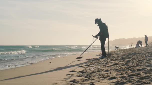 A man walks along the beach with a metal detector looking for luxury items. — ストック動画