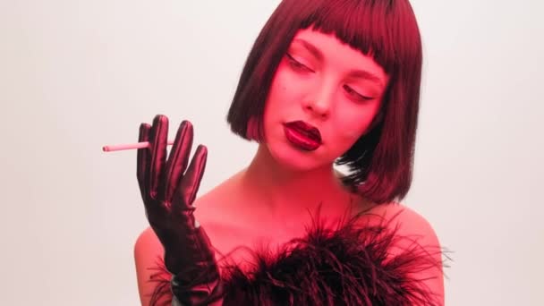 Ritratto di moda di una ragazza a luce rossa su uno sfondo bianco con una sigaretta in bocca. Labbra nere e pettinatura kare con frecce nere primo piano. — Video Stock