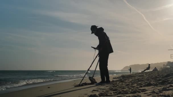 Un uomo cammina lungo la spiaggia con un metal detector alla ricerca di oggetti di lusso. — Video Stock