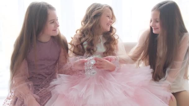 Trois amies sont assises sur le canapé et regardent la couronne de la princesse. Enfants heureux dans l'appartement sur un fond clair. — Video