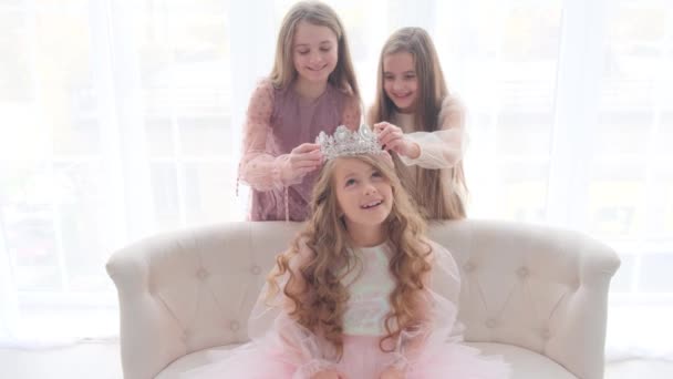 Trzy przyjaciółki siedzą na kanapie i patrzą na koronę księżniczki. Szczęśliwe dzieci w mieszkaniu na jasnym tle. — Wideo stockowe