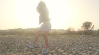 Kumsalda mutlu bir kız ayaklarıyla kum saçar ve tatilin tadını çıkarır. Yaz akşamı, gün batımı ve bir çocuğun siluetiyle..