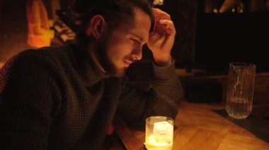 A young drunk man sits at the bar at the bar with a glass of whiskey.
