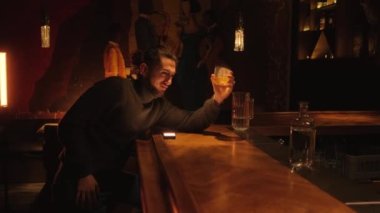 A young drunk man sits at the bar at the bar with a glass of whiskey.