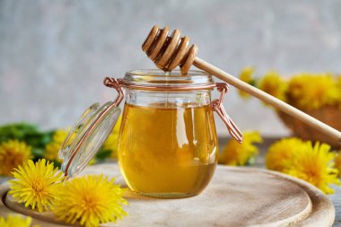 A glass jar of dandelion honey with a dipper and fresh flowers