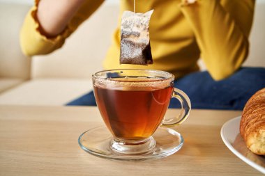 A tea bag is being lifted above the cup by a teenage girl, with a croissant in the foreground