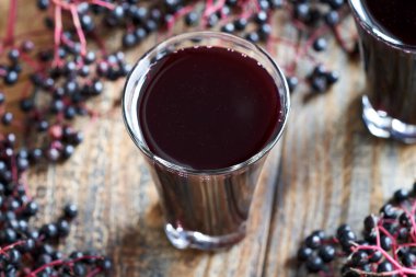 Bir bardak ev yapımı siyah yaşlı şurubu. Arka planda tahta bir masada taze mürver var.