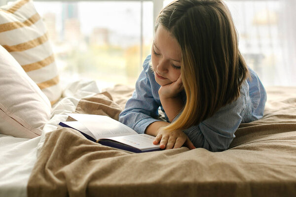 8-9 years old caucasian girl lying on cozy bed, reading a book. Concepts of home and comfort