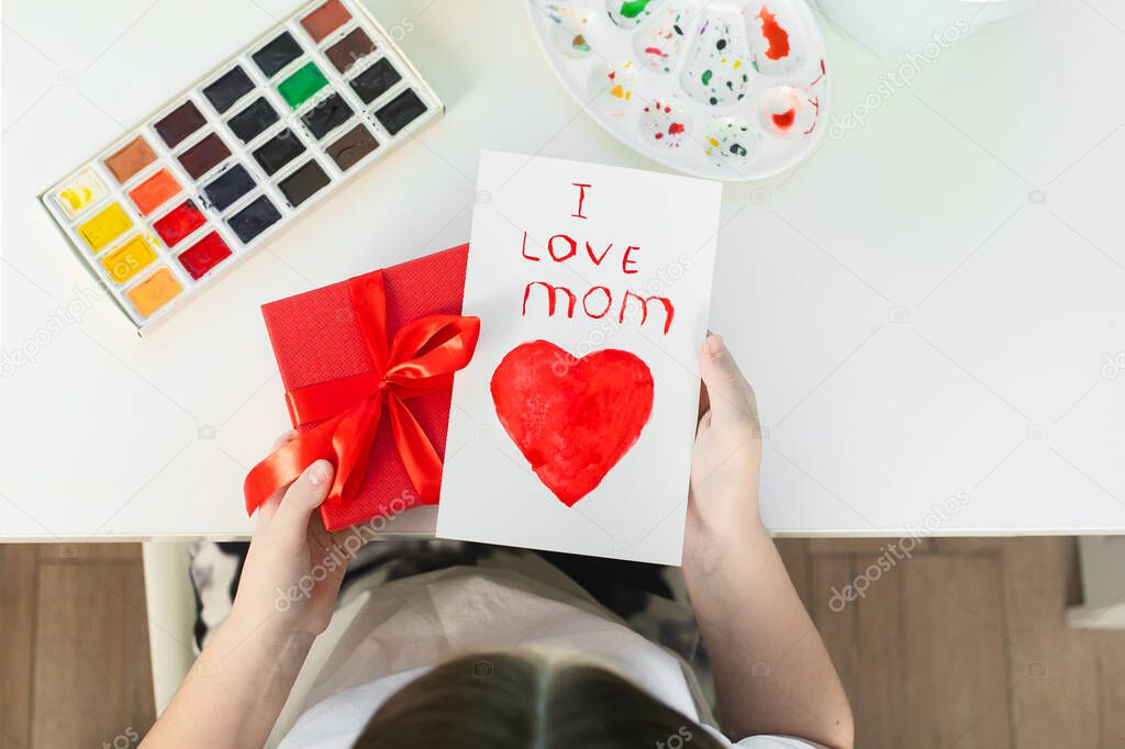 Girl holding in hands a red gift box and handmade greeting card for mom. Concept of Mother s day, woman s day.