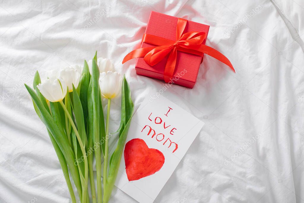 White tulips flowers, red gift box and greeting card for holiday on white bed. Top view, flat lay. Concept of womans day, mothers day