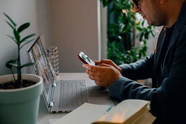 Focused European young man wearing glasses using laptop, texting on phone, working online on computer at home.