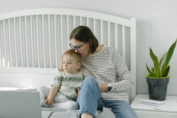 Young single mother mom with toddler boy son sitting on bed using laptop, searching web, watching cartoons together. Concept of work from home, freelancer