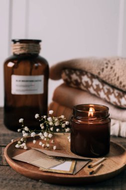 Burning candle in small amber glass jar, flowers of gypsophila and stack knitted seasin sweaters. Cozy lifestyle, hygge concept
