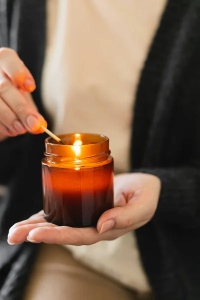 Woman holding in hand candle and light candle in small amber glass jar with wooden wick. Zen and relax concept.