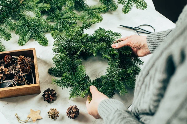 Woman in gray knitted sweater making a christmas fir wreath. Christmas decorating for home interior.
