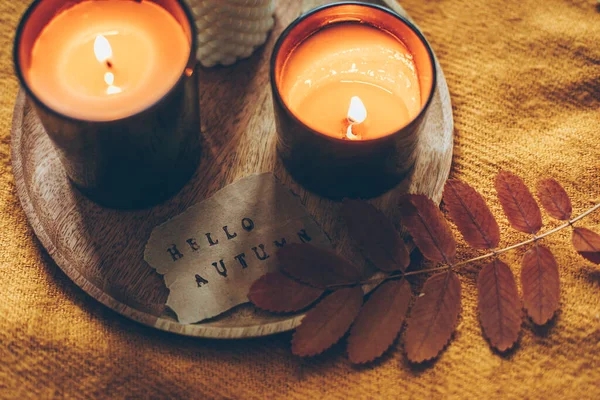 Wooden tray with burning candles and card with text hello autumn on orange wool plaid on bed. Aututmn mood, hygge, cozy concept