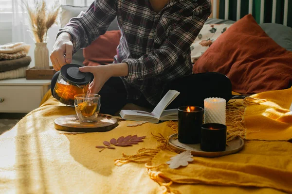 Young woman pouring a tea while sitting on bed, reading book, relaxing at home in autumn fall season. Concept of cozy autumn at home, hygge