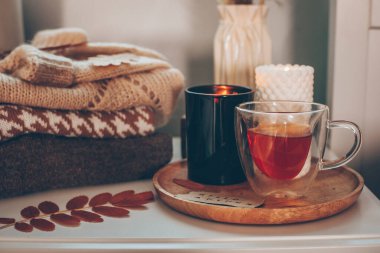 Still life details. Cup of tea and candles on wooden tray and stack of warm woolen sweaters. Autumn mood, cozy home, hygge concept