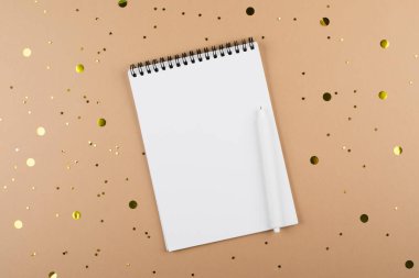 Top view of white spiral notebook note pad and pen on beige background with golden confetti. Mockup concept. Copy space.