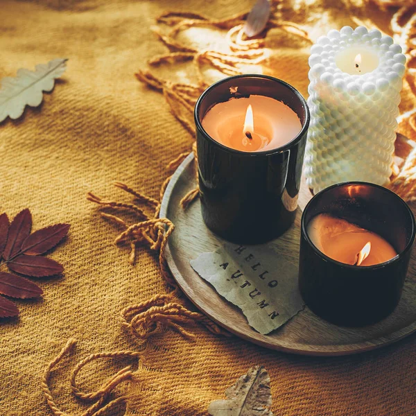 Wooden tray with burning candles and card with text hello autumn on orange wool plaid on bed. Autumn mood, hygge, cozy concept