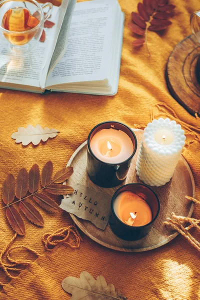 Burning candles with card hello autumn, herbal tea in glass cup and open book on yellow plaid. Reading, relaxation, recreation at home. Autumn mood. Top view