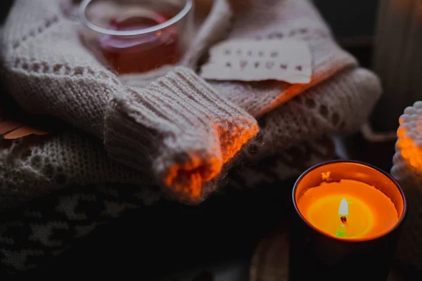 Two burning candles, glass cup of tea and stack of warm cozy sweaters om bedtable. Cozy autumn evening at home. Selective focus