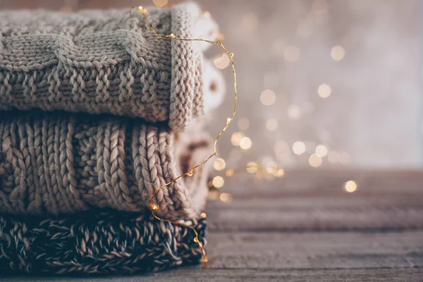 Stack of cozy winter or autumn knitted sweaters and garland lights on wooden background. Warm Cozy Concept. Copy space