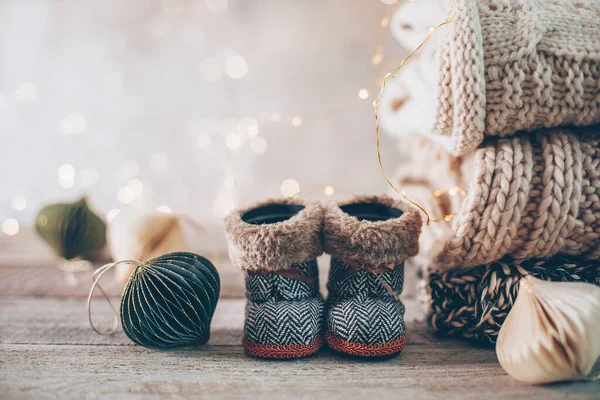 Stack of cozy winter knitted sweaters, cute small boots and christmas ornaments on a blurred background with bokeh. Warm Cozy Concept. Copy space