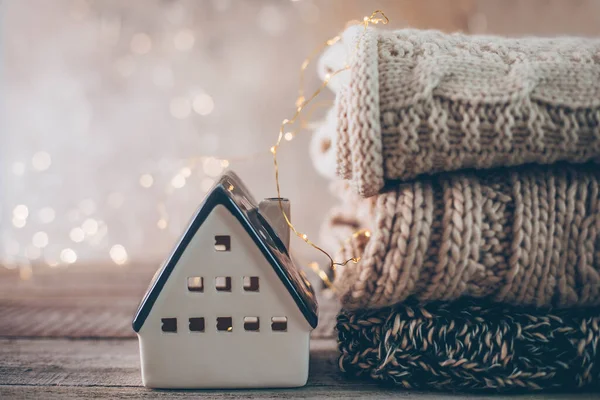 Stack of cozy winter knitted sweaters, ceramic house on a blurred background with bokeh. Warm Cozy Concept.