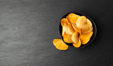 Potato chips pile in black bowl on dark stone background closeup. Crispy thin potato snack heap, fast food snacks with greens top view