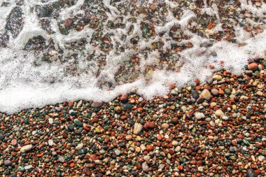 Sea waves on colored pebble beach texture background. Transparent ocean water, multicolored rocky shore pattern, summer mockup with copy space, stone shoreline, vacation concept