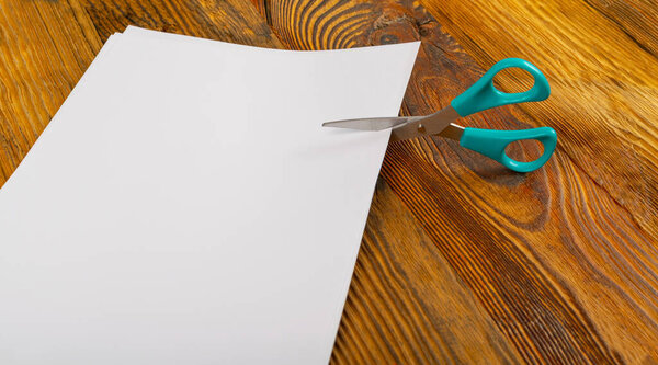 Scissors cutting folded paper. Blue scissors cuts white paper piece on wooden background side view