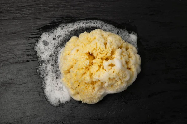 Natural sea sponge with soap foam on black background top view. Yellow sponge, eco body care concept, eco friendly hygiene accessory