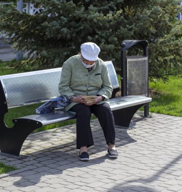 Novomoskovsk, Ukraine - May 13, 2021: elderly woman in mask on bench on street in city reads book