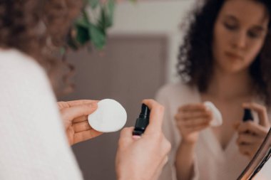 Aynada kendine bakım yapan genç bir kadın, cilt için aroma yağı ya da serum kullanıyor.