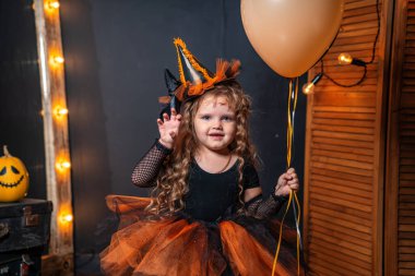 Mutlu Cadılar Bayramı. Arka plan dekorasyonunda balkabağı olan sevimli küçük cadı. Çocuk Cadılar Bayramı için hazırlanıyor. Bebek elinde balkabağı şeklindeki bir fenerle korkar. Şaka mı şeker mi?