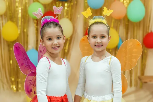 Two little girls in carnival costumes of fairy butterflies are standing on a shiny gold background with balloons. Children\'s theater. A dance group.