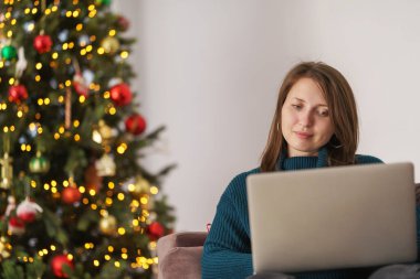 Genç bir kadın, Noel 'de rahat bir yerde dizüstü bilgisayarla alışveriş yapıyor. Kız kanepede oturuyor. Arka planda süslü bir Noel ağacı var. Kış satışlarından hediyeler alıyorum..
