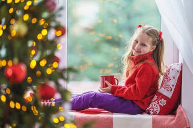 Küçük tatlı bir kız elinde bir fincan sıcak içecekle pencerenin yanında oturuyor. Madalyalı Noel ağacı. Sıcak giysili, battaniyeli ve penceresinde yastığı olan bir çocuk. Mutlu Noeller ve mutlu Noeller..