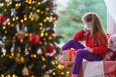 Sevimli küçük kız elinde hediye kutusuyla pencerenin kenarında oturuyor. Dekore edilmiş Noel ağacı, battaniyeli ve penceresinin yanında yastıklı çocuk. Çocuğun yüzünde maske. Mutsuz çocuk karantinada.