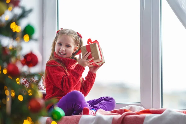 一个可爱的小女孩带着礼品盒坐在窗边 装饰过的圣诞树一个穿着保暖衣服 带着毛毯和枕头的孩子在窗边 圣诞快乐 节日快乐 — 图库照片