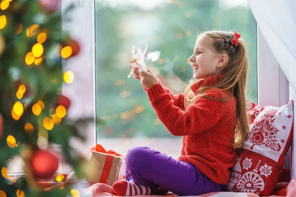 一个可爱的小女孩和一个圣诞树玩具坐在窗边 装饰过的圣诞树一个穿着保暖衣服 带着毛毯和枕头的孩子在窗边 圣诞快乐 节日快乐 — 图库照片