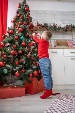 Mutlu noeller. Şirin küçük kız evdeki Noel ağacını süslüyor. Geyik boynuzlu beyaz kazaklı çocuk, elinde hediyeyle yerde oturuyor ve Noel ağacındaki oyuncağı ayarlıyor..