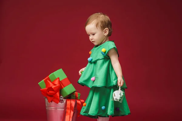 身穿圣诞树装束 头戴喜庆饰带的小女孩站在红色背景的桶里 一个孩子手里拿着圣诞玩具 — 图库照片