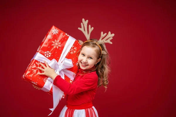 快乐的小女孩4岁在驯鹿鹿角在明亮的红色背景 孩子很高兴 手里拿着礼物 把它向前拉了拉 送礼物新年销售 — 图库照片