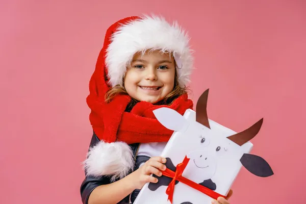 牛の形をしたクリスマスのギフトボックスとサンタの帽子の幸せな小さな笑顔の女の子 子供はピンクの背景でただ笑う — ストック写真